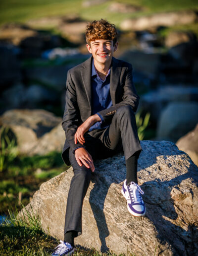 Bar Mitzvah boy wearing a blazer and dress pants, smiling while sitting on a stone outdoors at Union League Liberty Hill, captured by PCSI Studio – elegant and natural Mitzvah portrait photography.