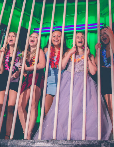 Group of girl friends smiling with the Bat mitzvah girl in her purple beaded gown looking like they're in Jail behind bars at Beat Street in Huntingdon Valley, Pennsylvania.