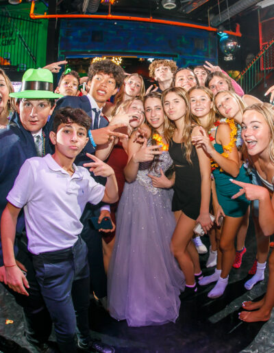 Bat Mitzvah girl in her purple beaded gown in a crowd of friends giving the peace sign wearing tropical colored hats and leis at Beat Street Huntingdon Valley, PA