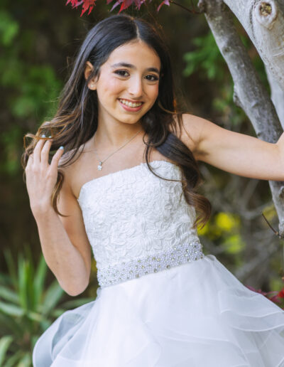 Bat Mitzvah girl in a short white ruffle dress with pearl and sequin belt smiles beside a tree at Philmont Country Club in Huntingdon Valley, Pennsylvania.