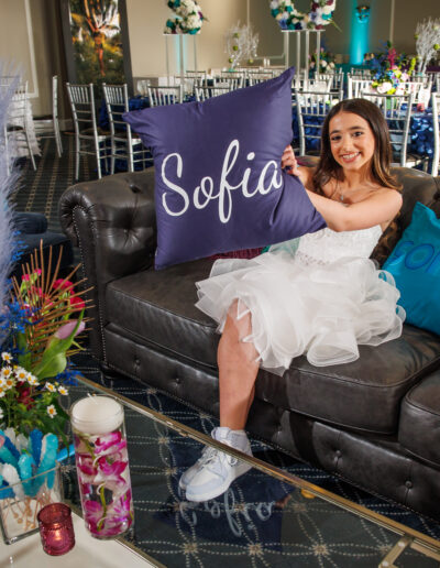 Bat mitzvah girl holding a personalized pillow with her name amid vibrant blue, purple, and green peacock-inspired decorations at Philmont Country Club in Huntingdon Valley Pennsylvania, photographed by PCSI PHOTO.