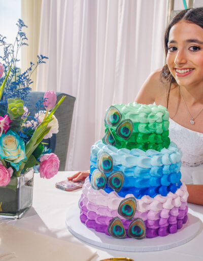 Bat mitzvah girl with her colorful peacock-themed tiered cake decorated in blue, green, and purple hues at Philmont Country Club in Philadelphia, photographed by PCSI PHOTO.