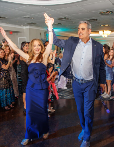 Bat Mitzvah girl's parents, mom in a navy gown and dad in a blue suit raise their arms and smile as guests cheer during their grand entrance at the celebration at Philmont Country Club in Huntingdon Valley Pennsylvania