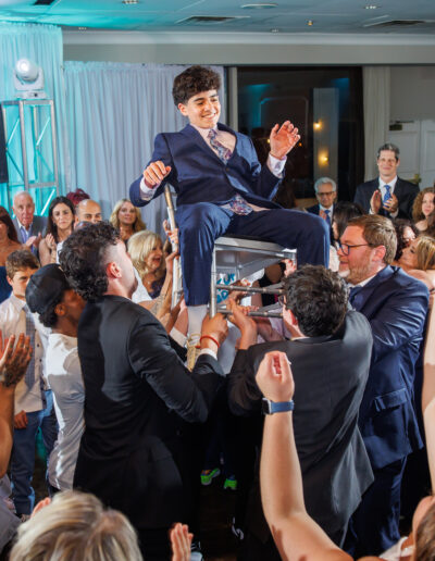Bat Mitzvah Girl younger brother dressed in Blue suit celebrating as the crowd lifts him up in a chair under vibrant blue lights during the hora dance at Philmont Country Club in Huntingdon Valley, Pennsylvania.