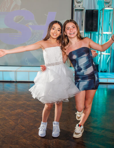 Bat Mitzvah girl in white ruffle dress and her friend dressed in a blue tye dye dress laughing together on the blue and lavender-lit dance floor at Philmont Country Club in Huntingdon Valley, Pennsylvania.