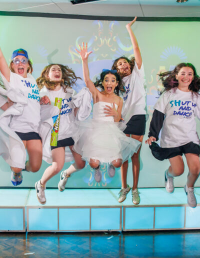 Bat Mitzvah girl in her strapless white ruffle dress jumps with friends on stage, illuminated by blue and lavender lights during her celebration at Philmont Country Club, Huntingdon Valley, Pennsylvania