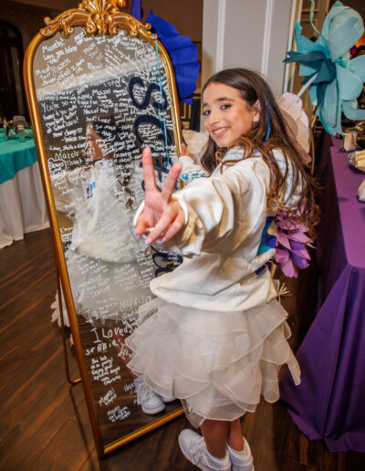 Bat Mitzvah girl wearing her white ruffle cocktail dress flashes a peace sign while signing a gold-framed mirror guest board at Philmont Country Club in Huntingdon Valley, Pennsylvania.