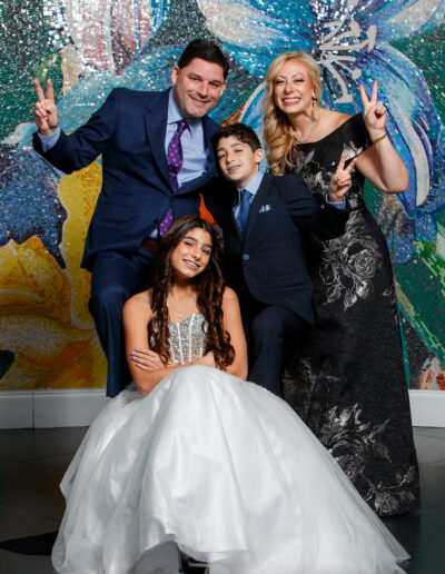 Bat Mitzvah girl in a white beaded gown kneeling in front of her family giving peace signs with big smiles in front of a colorful wall at Vie by Cescaphe in Philadelphia
