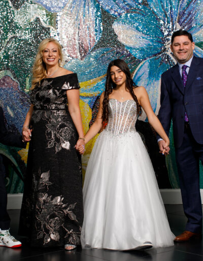 Bat Mitzvah girl in a white beaded strapless gown holding hands with her family in navy and light blue in front of a colorful wall at vie by Cescaphe in Philly