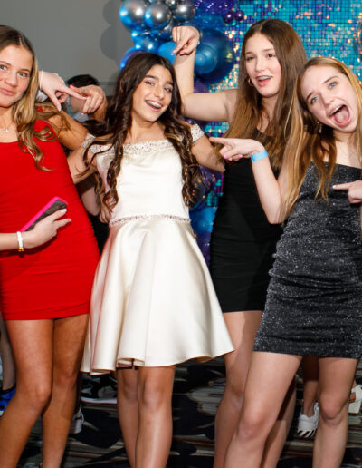 Bat mitzvah girl in a champagne colored beaded dress beaming surrounded by her best girlfriends pointing towards her at Vie by Cescaphe Philly