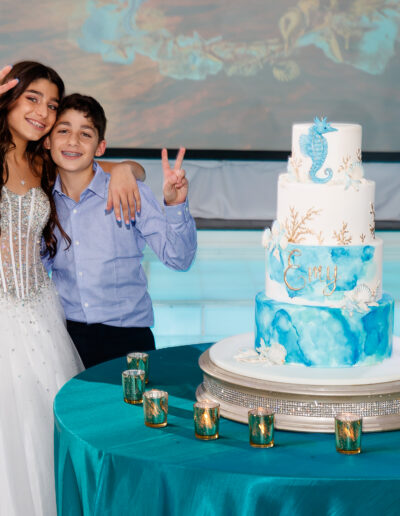 Bat Mitzvah girl in her stunning white embellished gown alongside her little brother giving peace signs beside the white and turquoise sea life cake adorned with starfish, coral and seashells at Vie by Cescaphe in Philly