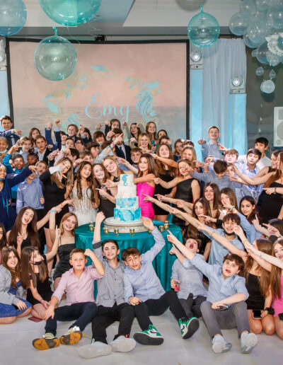 Bat Mitzvah girl in her white beaded gown surrounded by all her friends posing playfully on the dance floor with an ocean inspired 4 teired cake in whaite and turquoiseadorned with a seahorse and coral at vie by Cescaphe in Philadelphia