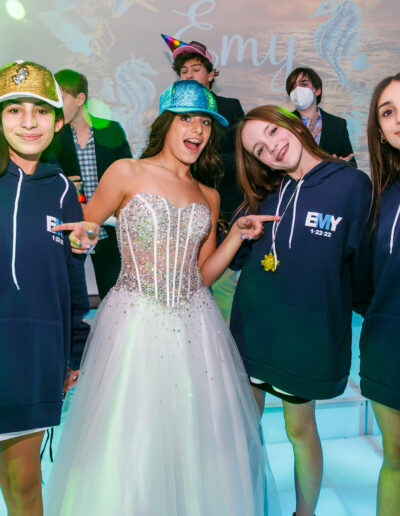 Bat Mitzvah girl in a white beaded tulle gown posing with friends wearing custom navy hoodies and baseball hats during celebration at Vie by Cescaphe in Philadelphia