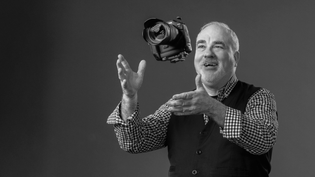 professional mitzvah photographer wearing a black vest and plaid shirt tossing a camera into the air