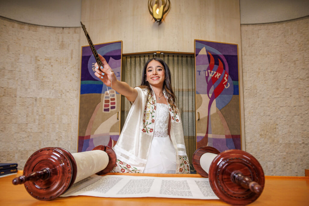 bat mitzvah girl smiling and holding yad while reading from the torah at shir ami synagogue
