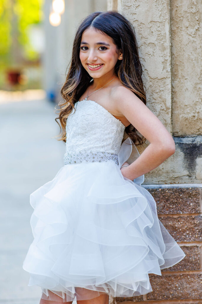 bat mitzvah girl wearing a white dress posing against a wall during outdoor photos at shir ami